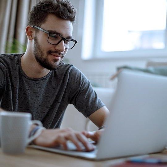 Man sitting at a table with laptop and cup of coffee staying connected with Viasat's satellite internet and the FCC's 负担得起的连接计划.